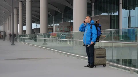 Passenger with suitcase and backpack, talking with phone on the airport terminal Stock-Footage 99016599