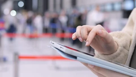 Passenger uses digital device amid airport crowd, seated near barriers 스톡 동영상 345942528