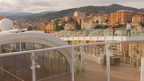 Passengers Stroll on Upper Deck of Cruise Ship Docked at Savona with City View Видео 335883653