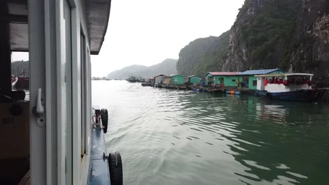 Passing an exotic, floating village in a boat on a cloudy day, Halong Bay Stock Footage