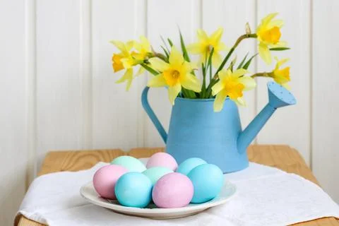 Pastel Easter eggs on a plate with yellow daffodils in a blue watering can,.. Stock Photos