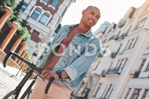 Pastime. Young guy walking on city street with bicycle looking aside smiling Stock Photos