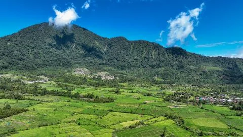 Patchwork fields and tiny rooftops cling to the foot of a towering jungle ridge Stock Photos