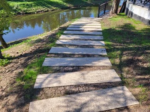 A path of concrete rectangles is laid along a shady spring lawn along the riv Stock Photos