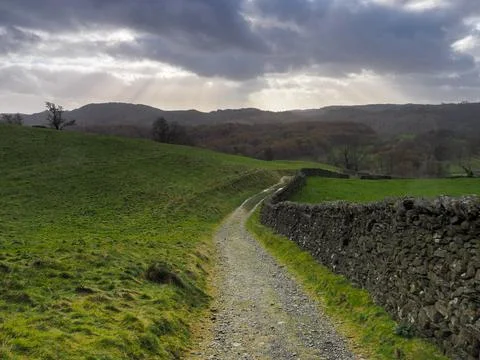 Path by dry-stone wall with rays of sunlight in a stormy sky, Lake District Stock Photos