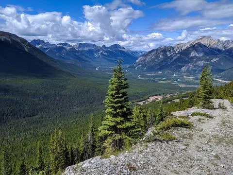 A Path Leads Down into a Valley Surrounded by Mountains and Forest Stock Photos