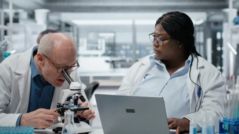 Pathologist examining biopsy slide while coworker documents findings on laptop Stock Footage 327244584