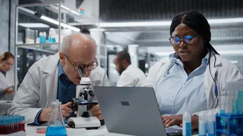 Pathologist examining biopsy slide while coworker documents findings on laptop Stock Photos