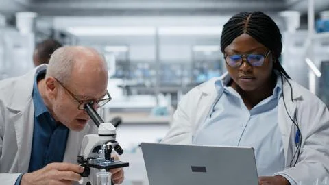 Pathologist examining biopsy slide while coworker documents findings on laptop Stock Photos