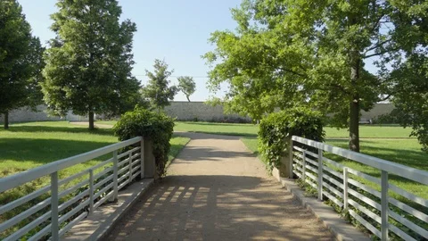 A pathway in a green park with trees and bushes over a bridge with white fence. 스톡 동영상 122767927