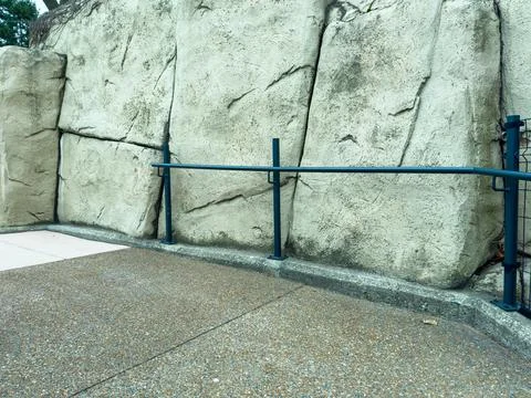 A pathway shows a concrete surface next to a large stone wall. A blue railing Stock-Fotos