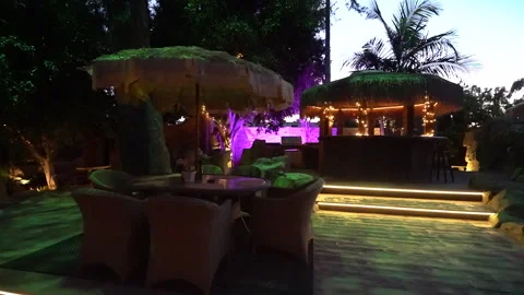 Patio dining set under thatched umbrella near a lit tiki bar Stock Footage 345538612
