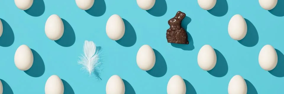 Pattern made of white eggs, chocolate banny and bird feather on blue backgr.. Stock Photos