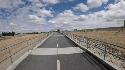 A paved multi-use bike path leads uphill along metal guardrails under a cloudy s Vídeos de archivo 348096278
