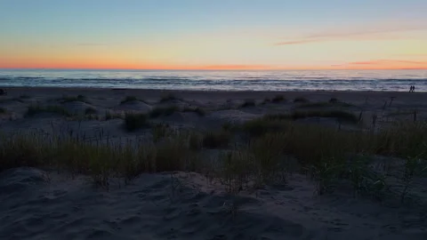Peaceful sunset over empty sandy beach with dune grass, calm ocean horizon, and Stock Footage 338134692