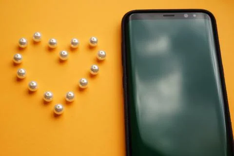 Pearl beads on an orange table in the shape of a heart. Black smartphone in a Stock Photos