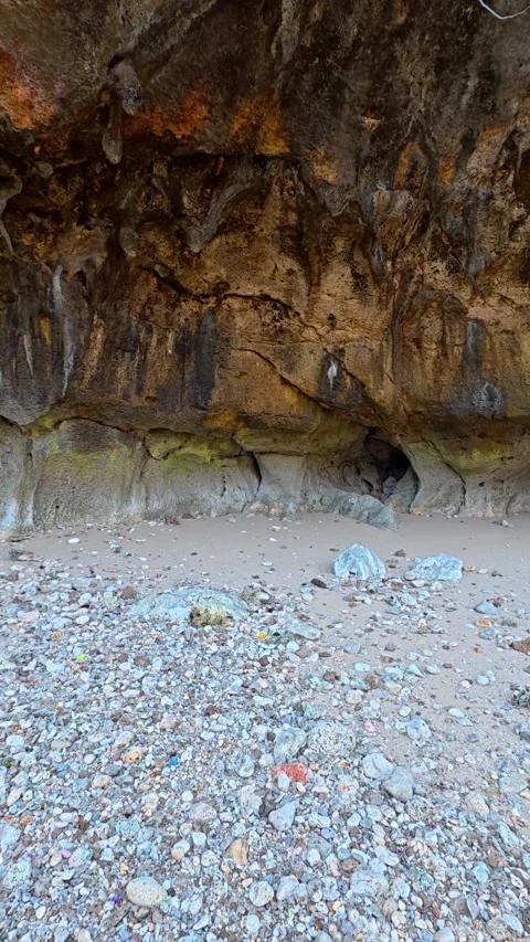 Pebble beach beneath rugged coastal cave and weathered rock cliff Stock Footage 346739444