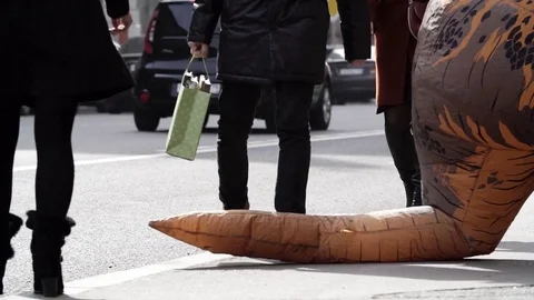 Pedastrians steps over tail of dinosaur costume mascot on city sidewalk Vidéo 84928953