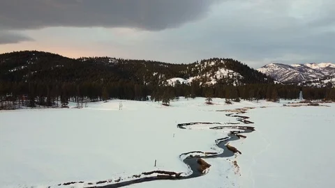 Pedestal down shot over vast snow covered field with winding creek, forest an Stock Footage 125384587