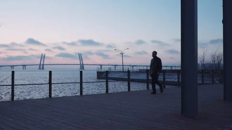 Pedestrian hastens down seafront walkway, majestic bridge stretching across view Stock Footage 270799660