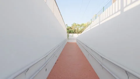 A pedestrian ramp with pink flooring between white walls and greenery, Cote Stock Footage 344799496