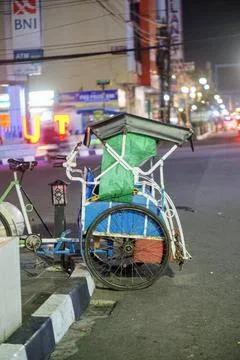 A pedicab is parked on the side of a city road at night with the lights of busy Stock Photos