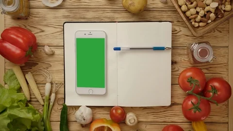 Pen rolling on blank notebook with smartphone on kitchen table with vegetables Vidéo 121585810