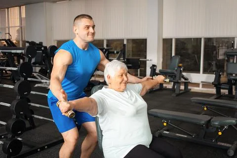 A pensioner performs an exercise with dumbbells on simulator in gym. coach sh Stock Photos