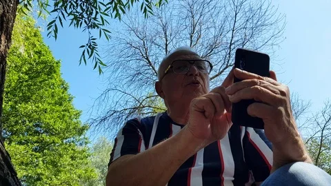Pensioner sits texting on smartphone at tree in summer park Stock Footage 112396133