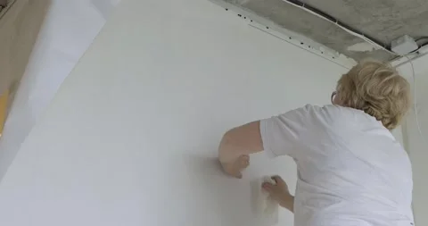 Pensioner work part-time in construction team, glues wallpaper on prepared walls Stock Footage 131034798