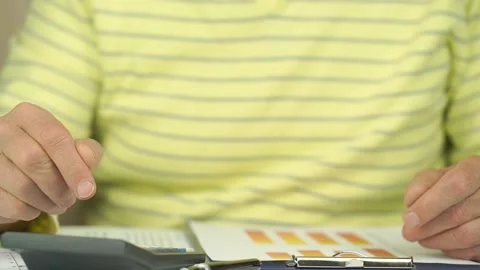 Pensioner in yellow pullover counts on calculator and writes Video stock 134239815