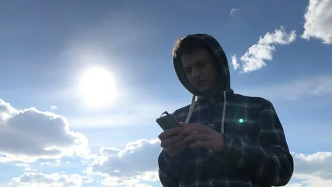 Pensive boy in hood using smartphone outdoors on the cloudy sky background. Slow Vidéo 106636759