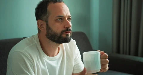 A pensive brunette guy with a beard in a white T-shirt who drinks hot tea from a Stock Footage 280669797
