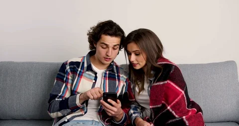 Pensive couple sitting on sofa, using smartphones, smiling and looking at each Stock Footage 125614860
