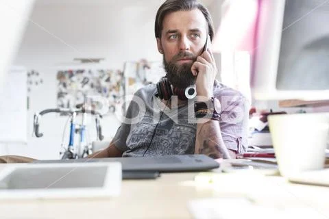 Pensive design professional with headphones talking on cell phone at desk Stock Photos