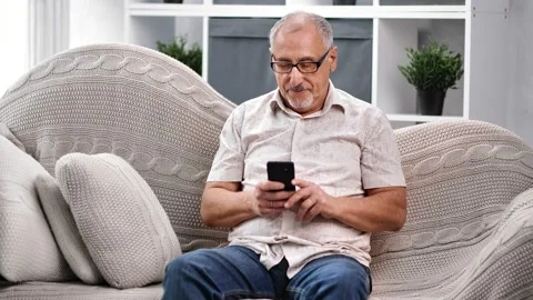 Pensive elderly 60s man smiling chatting use smartphone sit on couch. Medium Stock Footage 139559831