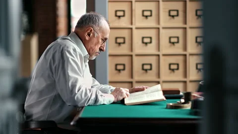 Pensive elderly man reading text paper book sitting at vintage desk table home Stock Footage 165036529