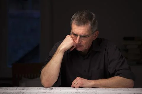 Pensive engineer or architect in dark shirt looking at blueprint at home by n Stock Photos