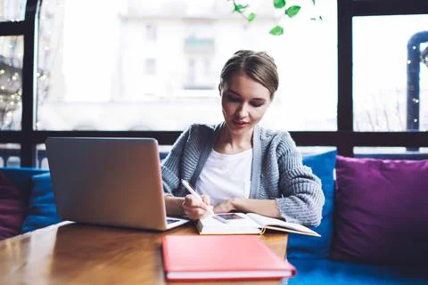 Pensive freelancer writing notes in notebook while working on laptop in cafe Stock Photos