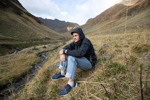 Pensive guy in a black jacket with a hood sits on the grass in the mountains Stock Photos