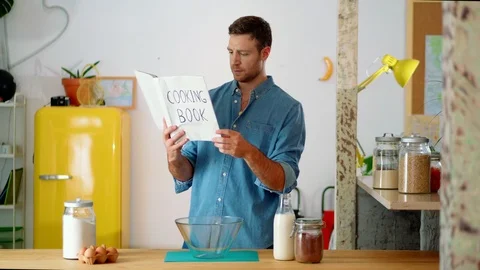 Pensive guy standing at kitchen holding cooking book confused with culinary Stock Footage 114915537