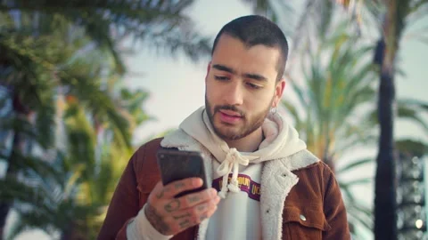 Pensive handsome Hispanic man using smartphone device outdoors palm trees on Stock Footage 136673727