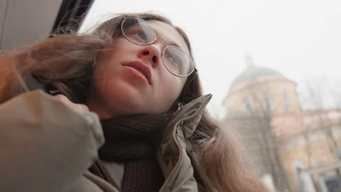 Pensive lady in coat views rainy cityscape through window. Quiet woman with Stock Footage 345539429