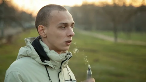 Pensive man smoking electronic cigarette and exhale clounds of vapor in the park Vidéo 121061753