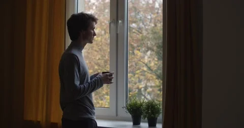 Pensive man standing by window indoors holding cup of tea slow motion. Back side Stock Footage 142983121
