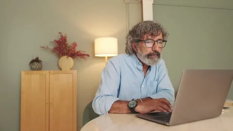 Pensive middle-aged man in glasses sitting in the living room working on laptop Stock Footage 254913182