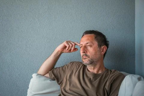 Pensive middle-aged man sits on a couch against a gray wall, fingers to his.. Stock Photos