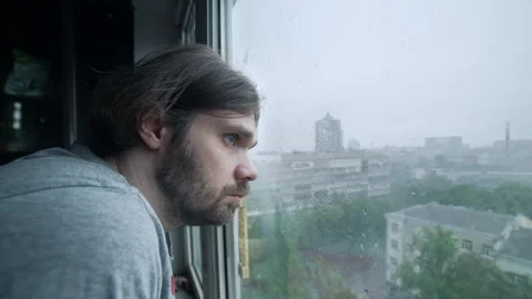 Pensive sad man stands at the window on rainy day at view of city skyline 스톡 동영상 277646158