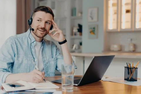 Pensive thoughtful man makes notes uses laptop computer headset writes down Stock Photos
