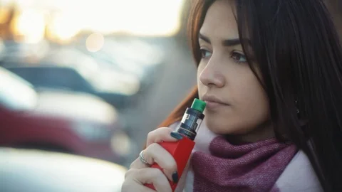 Pensive woman vaping e-cigarette between  cars on the parking zone near the Stock Footage 121482998